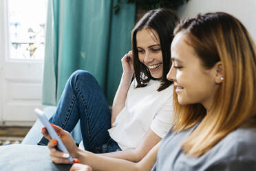 Smiling women sharing mobile phone at home - XLGF01759