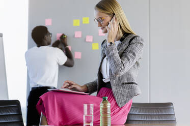 Businesswoman talking on smart phone while male colleague working in background at office - BMOF00573