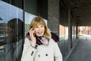 Smiling woman talking on smart phone while leaning on glass - IHF00443