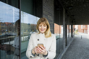 Mature woman using smart phone while standing by glass - IHF00444