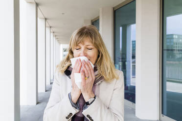 Mature woman sneezing while standing at corridor - IHF00447