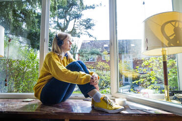 Woman wearing headphones listening music while sitting on window sill at home - IHF00453