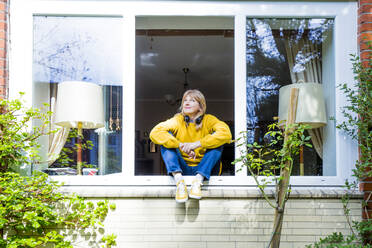 Reife Frau blickt nach oben, während sie zu Hause auf der Fensterbank sitzt - IHF00454