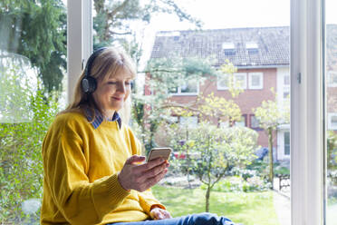 Ältere Frau hört Musik über Kopfhörer, während sie ihr Smartphone zu Hause benutzt - IHF00455
