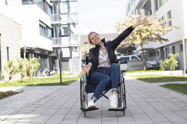 Glückliche behinderte Frau mit ausgestreckten Armen im Rollstuhl sitzend in einem Wohngebiet - SGF02819