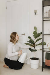 Schwangere Frau mit Gießkanne beim Betrachten einer Topfpflanze zu Hause - SMSF00537