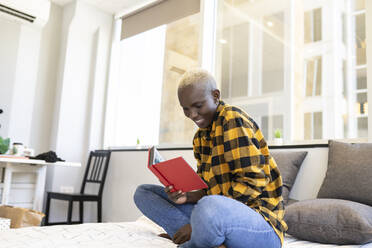 Smiling woman reading book while sitting on bed at bedroom - JCCMF02319