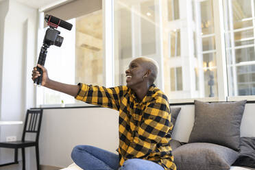 Smiling woman holding video camera while sitting on bed at home - JCCMF02320