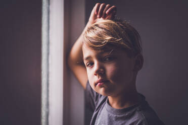 Junge mit haselnussbraunen Augen, der vor einem Fenster steht und in die Kamera schaut. - CAVF94054