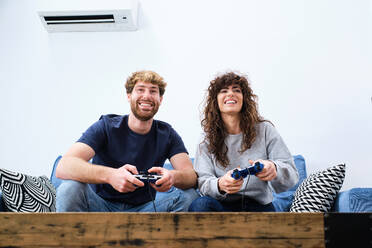 From below cheerful and excited young couple in casual clothes playing video game on console in stylish living room - ADSF24086