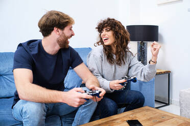 Side view of cheerful and excited young couple in casual clothes playing video game on console in stylish living room - ADSF24087