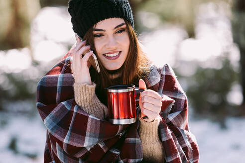 Fröhliche Frau in warmes Plaid gehüllt, auf einem Baumstamm im Winterwald sitzend und mit dem Handy telefonierend an einem sonnigen Tag - ADSF24099