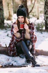 Fröhliche Frau in warmes Plaid gehüllt, die auf einem Baumstamm im Winterwald sitzt und an einem sonnigen Tag auf ihrem Mobiltelefon surft - ADSF24100