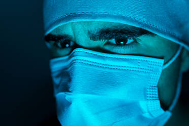 Closeup of male surgeon in medical mask looking away in dark room with blue neon light - ADSF24101