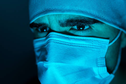 Nahaufnahme eines männlichen Chirurgen mit medizinischer Maske, der in einem dunklen Raum mit blauem Neonlicht wegschaut - ADSF24101