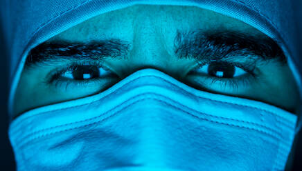 Closeup of male surgeon in medical mask looking at camera in dark room with blue neon light - ADSF24102