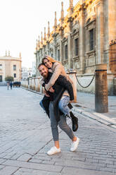 Playful man piggybacking cheerful woman while having fun in city street and looking at camera - ADSF24125