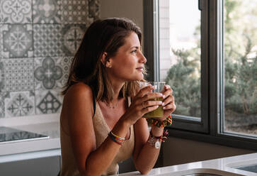 Caucasian girl is having a green vegetable juice in front of her big kitchen window - ADSF24135