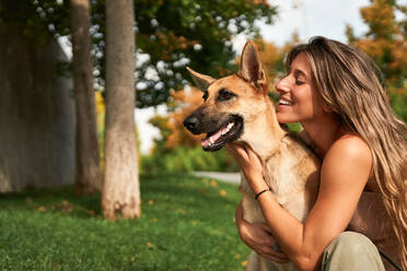 Positive Besitzerin, die ihren Deutschen Schäferhund umarmt, während sie zusammen auf dem Rasen im Park sitzen - ADSF24143