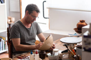 Man sitting at table with brushes and drawing sketches on handmade ceramic plate - ADSF24154