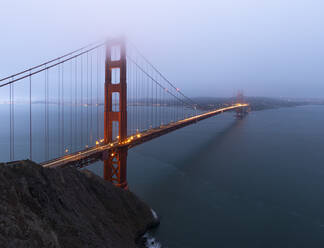 Golden Gate Bridge mit leuchtenden Lichtern über ruhigem Meer gegen Küstenstadt mit Nebel auf Abendzeit in San Francisco bedeckt - ADSF24158