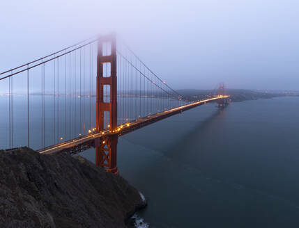 Golden Gate Bridge mit leuchtenden Lichtern über ruhigem Meer gegen Küstenstadt mit Nebel auf Abendzeit in San Francisco bedeckt - ADSF24158