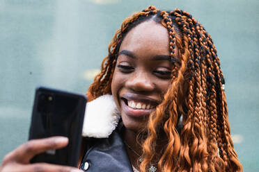 Fröhliche Afroamerikanerin in modischem Outfit und mit Zöpfen, die in der Nähe einer Glaswand eines Gebäudes steht und auf ihrem Smartphone in der Stadt surft - ADSF24159