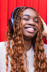 Positive African American female in wireless headphones enjoying songs with closed eyes while listening to music on street against red wall - ADSF24160