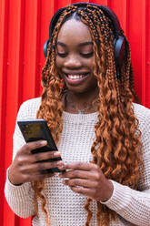 Positive afroamerikanische Frau mit kabellosen Kopfhörern, die auf der Straße vor einer roten Wand Musik hört - ADSF24164