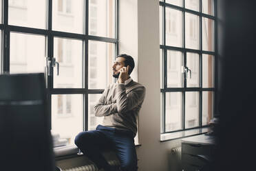 Männlicher Unternehmer, der am Fenster in einem kreativen Büro mit seinem Smartphone telefoniert - MASF23158