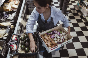 Hohe Winkelansicht der Verkäuferin, die Obst und Gemüse in einer Kiste im Delikatessengeschäft anordnet - MASF23213