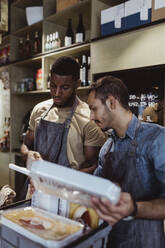 Male colleagues holding package food while standing at delicatessen shop - MASF23224