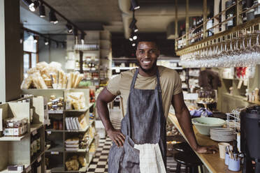 Porträt eines lächelnden männlichen Inhabers mit Hand auf der Hüfte in einem Feinkostladen - MASF23225