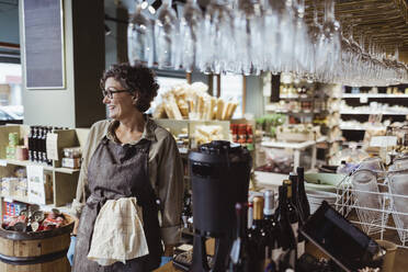 Happy female owner looking away while standing in deli store - MASF23230