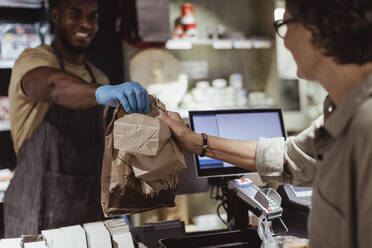 Male owner giving package order to female customer in delicatessen shop - MASF23242