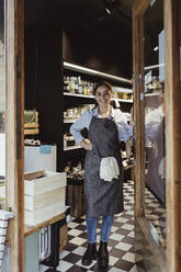 Portrait of smiling young female owner standing by door of delicatessen shop - MASF23265