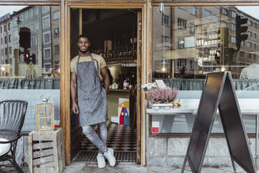 Smiling male owner leaning on doorway of delicatessen shop - MASF23275