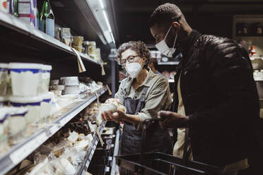 Female owner holding product while male customer standing by in deli store during COVID-19 - MASF23283