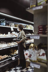 Female owner arranging food product on rack at deli shop during pandemic - MASF23296