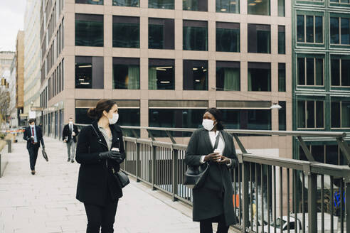 Female colleagues talking with each other while walking on bridge in city - MASF23340