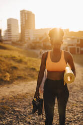 Junge Sportlerin mit Sportschuh und Trainingsmatte bei Sonnenuntergang an Land stehend - MASF23412