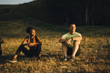 Male and female athletes looking away while sitting on grass during sunset - MASF23419