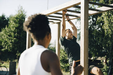 Male athlete hanging on monkey bar while sportswoman looking at him in park on sunny day - MASF23448