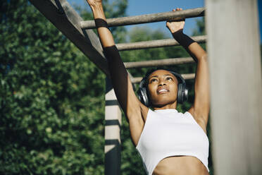 Sportswoman listening music through headphones while exercising on monkey bar in park - MASF23450