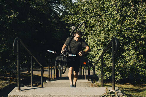 Aktiver männlicher Sportler, der im Park eine Treppe hinaufsteigt und ein Fahrrad trägt - MASF23456