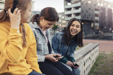 Glückliche Freundinnen, die ihr Smartphone benutzen, während sie sich an einer Stützmauer unterhalten - MASF23465