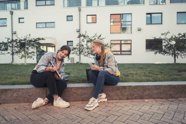 Teenage girl showing digital tablet to female friend while sitting on retaining wall against building - MASF23486