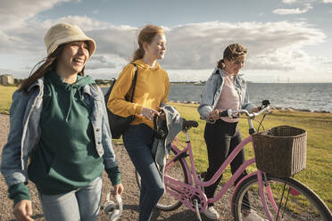 Happy female friends with bicycle walking at lakeshore against cloudy sky - MASF23494