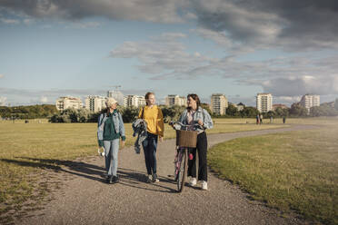 Teenage girl with bicycle walking by female friends in park - MASF23495