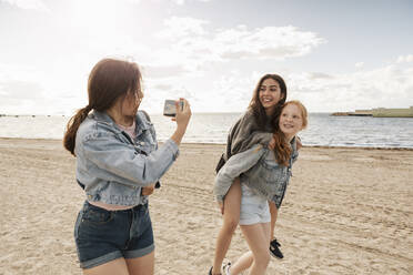 Teenager-Mädchen, das Huckepack reitet, während Freundinnen am Strand gegen Meer und Himmel fotografieren - MASF23507